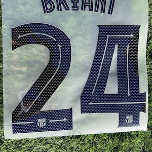 May include: A navy blue heat transfer vinyl sheet with the name "BRYANT" and the number "24" in a stylized font. The number has a dotted pattern and includes a crest at the bottom. The sheet is on a green grassy surface.
