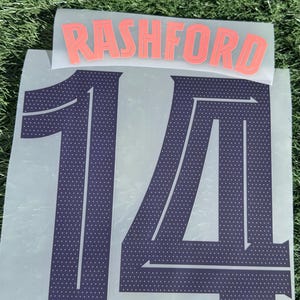 Pode incluir: Uma folha com o nome "RASHFORD" em letras coral acima do número "14" em azul escuro. O número tem um padrão pontilhado e um logotipo da equipe na parte inferior. O fundo é verde.