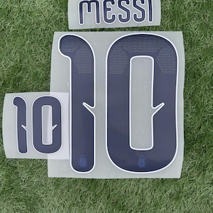 Puede incluir: Juego de números y nombre de transferencia de calor azul marino para una camiseta de fútbol. El número "10" se muestra en dos tamaños, junto con el nombre "MESSI" en una fuente a juego. El logotipo de la Asociación Argentina de Fútbol es visible.