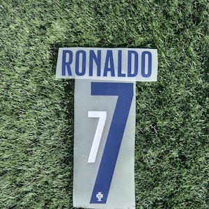 Puede incluir: Un letrero rectangular con el nombre "RONALDO" en letras azules sobre un letrero más grande con el número "7" en azul y blanco. El fondo es verde.