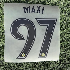 Puede incluir: Una calcomanía gris con el nombre "MAXI" encima del número "97" en negro. El número tiene un contorno blanco y pequeños logotipos en la parte inferior. La calcomanía está sobre una superficie de hierba verde.