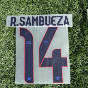 May include: A clear sheet with the name "R. SAMBUEZA" in black block letters above a large number "14". The number is navy blue with red trim and a small logo. The text "NUMERO OFICIAL 2015-2016" is also present.