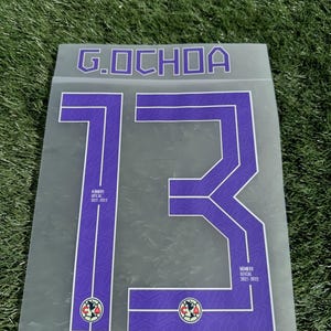 May include: A transparent sheet featuring the name "G. OCHOA" and the number "13" in purple with a white outline. The number includes the text "NUMERO OFICIAL 2021-2022" and a team logo. The background is green grass.