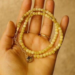 May include: A delicate necklace featuring small, square, yellow-green beads. The necklace has a gold chain and a small, round pendant with a patterned design. The necklace is displayed on a hand.