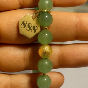 May include: A beaded bracelet featuring green and gold beads. The bracelet has a gold-colored charm with the number "888" on it. The beads are round and smooth, with a mix of green and gold colors.