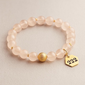 May include: A rose quartz beaded bracelet with a gold-colored charm. The bracelet features pale pink, round beads and a larger gold bead. The gold charm is a hexagon shape with the number "222" in black.