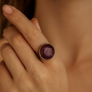 May include: A large, round, ruby-colored gemstone ring. The ring has a gold-colored band with intricate detailing. The gemstone is faceted, catching the light and displaying a deep red hue. The ring is worn on a finger.