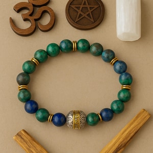 May include: A beaded bracelet featuring green and blue spherical beads, separated by gold-colored spacers. A larger, silver-colored bead with a geometric design is centered. The bracelet is displayed with spiritual and natural elements.
