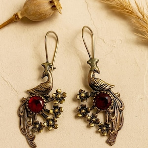 May include: A pair of antique-style earrings with a peacock motif. The earrings have a bronze finish and a large, round, red gemstone. The peacock's tail feathers and floral details enhance the design. The earrings are for pierced ears.