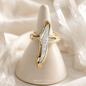 May include: A gold-toned ring featuring a cross-shaped inlay of iridescent white mother-of-pearl. The metal frame has an organic, irregular edge surrounding the central stone. The ring is displayed on a smooth, beige conical stand.
