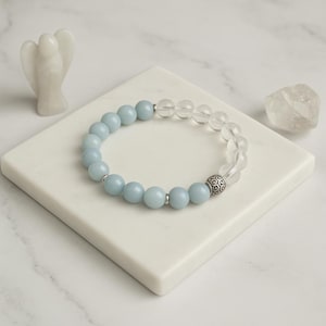 May include: A beaded bracelet featuring light blue and clear crystal beads. The bracelet has a silver-colored decorative bead and is displayed on a white square surface. A small white angel figurine and a clear crystal are also present.