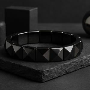 Op de afbeelding: Een zwarte armband met geometrische, piramidevormige kralen. De armband is gemaakt van gepolijst, donker materiaal en ligt op een donkere, getextureerde ondergrond. Een stijlvol accessoire.