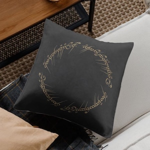 Puede incluir: Un cojín cuadrado negro con letras doradas en un diseño circular. El cojín está sobre un sofá blanco, con una mesa de madera y una botella de vidrio decorativa en el fondo. El diseño está inspirado en una novela de fantasía.