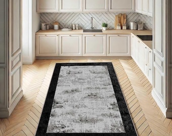 May include: A rectangular rug featuring a black border and a grey and white abstract design. The rug is positioned on a wooden floor within a kitchen, with white cabinets and a sink in the background.