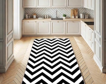 May include: A black and white chevron patterned rug in a kitchen setting. The rug is rectangular and covers a portion of the wooden floor. The kitchen features white cabinets and a light-colored countertop.