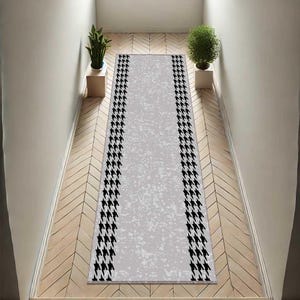May include: A rectangular grey runner rug featuring a black houndstooth pattern along its borders. The rug is laid on a wooden floor, framed by white walls. Two potted plants are positioned on either side of the rug.