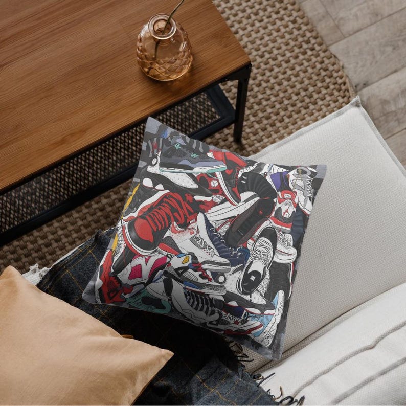 May include: Square throw pillow featuring a colorful collage of sneakers in various styles and colors, including red, black, white, and blue. The pillow is placed on a couch with other pillows and a wooden table.