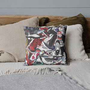 May include: Square throw pillow featuring a collage of colorful sneakers in various styles and colors, including red, black, white, and blue. The pillow has a gray border and is placed on a bed with neutral-toned pillows.