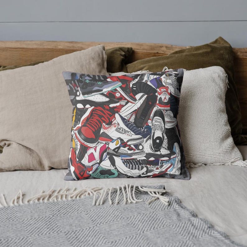 May include: Square throw pillow featuring a collage of colorful sneakers in various styles and colors, including red, black, white, and blue. The pillow has a gray border and is placed on a bed with neutral-toned pillows.
