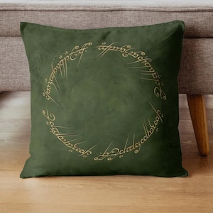 May include: A square, dark green pillow with a gold-colored inscription in a circular pattern. The inscription appears to be in a stylized script. The pillow is resting on a light gray surface.