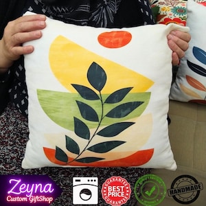 May include: White square pillow with an abstract design featuring yellow, green, and orange geometric shapes and a dark green leaf branch. The pillow has the text "Zeyna Custom GiftShop" and several icons.