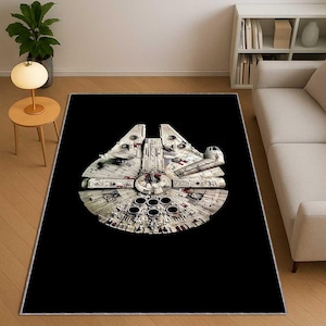 Puede incluir: Una alfombra rectangular negra con una imagen detallada de la nave espacial Millennium Falcon. La nave espacial es principalmente blanca y gris con detalles rojos y negros, sobre un fondo negro liso. La alfombra está diseñada para uso en interiores.