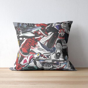 May include: Square throw pillow featuring a colorful illustration of various sneakers. The design includes red, black, white, and gray shoes, creating a collage effect. The pillow is placed on a wooden surface.
