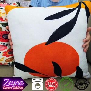 May include: Square decorative pillow with a white background, featuring an orange circle and black leaf design. The pillow has a yellow border. The pillow is being held by a person.