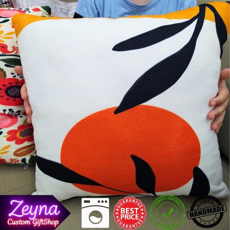 May include: Square decorative pillow with a white background, featuring an orange circle and black leaf design. The pillow has a yellow border. The pillow is being held by a person.
