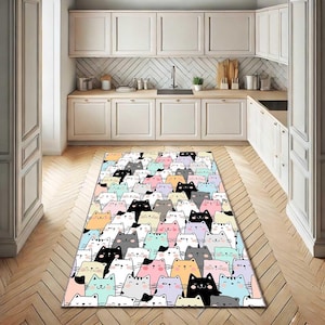 May include: A rectangular rug with a repeating pattern of cartoon cats in various colours, including black, white, pink, and blue. The rug is placed on a wooden floor in a kitchen setting, with white cabinets and a sink in the background.