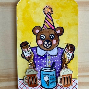 May include: Hand-painted earrings featuring a brown bear wearing a party hat and holding root beer bottles. The bear is on a yellow background with a blue ice cream cup and two beer mugs. The earrings are on a white card with a red and white checkered pattern.