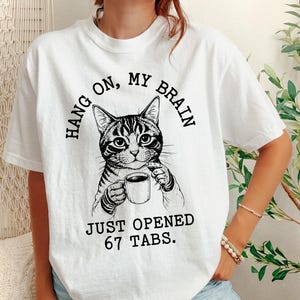 Può includere: T-shirt bianco con grafica in bianco e nero di un gatto che tiene una tazza di caffè. Il testo recita "HANG ON, MY BRAIN" sopra il gatto e "JUST OPENED 67 TABS" sotto. Stile casual e unisex.