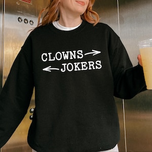 May include: Black sweatshirt with the text "CLOWNS -> JOKERS" in white. The sweatshirt has a crew neck and long sleeves. The word "Black" is displayed in a black rectangle at the bottom of the image.