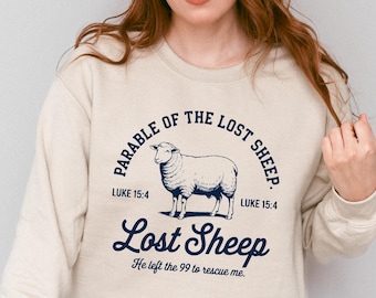 Può includere: Felpa color sabbia con grafica blu navy. La grafica presenta un'illustrazione di pecora e il testo "Parable of the Lost Sheep" e "Lost Sheep". È incluso anche il testo "Luke 15:4". La frase "He left the 99 to rescue me" è sotto.