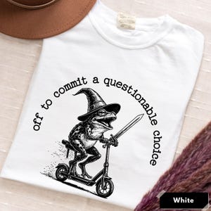 Könnte beinhalten: Weißes T-Shirt mit einer schwarz-weißen Grafik einer Frosch-Illustration, die einen Hexenhut trägt und ein Schwert hält, während es Roller fährt. Der Text "off to commit a questionable choice" ist um das Bild gedruckt.