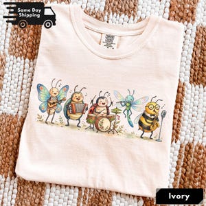 May include: A light ivory t-shirt featuring a colourful graphic of five anthropomorphic insects playing musical instruments, including a butterfly with a guitar, a beetle with an accordion, a ladybug on drums, a dragonfly with a flute, and a bee with a saxophone.