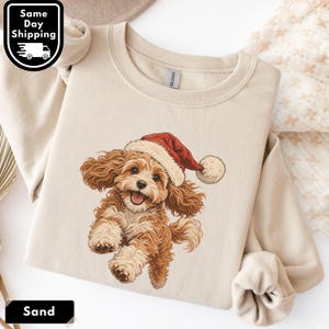 May include: A sand-colored crewneck sweatshirt featuring a graphic illustration of a fluffy brown puppy wearing a red and white Santa hat. The puppy is depicted in a playful, jumping pose with its mouth open, showcasing a cheerful expression.