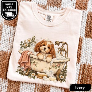May include: An ivory t-shirt featuring a graphic of a brown and white puppy sitting in a vintage clawfoot bathtub filled with bubbles. The design includes floral accents, a wooden brush, a bar of soap, and a gold-toned shower fixture.