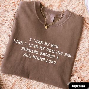 May include: Espresso-colored t-shirt with the text "I LIKE MY MEN LIKE I LIKE MY CEILING FAN RUNNING SMOOTH & ALL NIGHT LONG" in white. A gold-colored chain necklace with a pendant rests on the shirt.