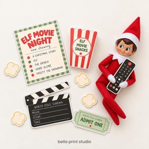 以下が含まれることがあります： 黒いリモコンを持つ赤と白のエルフの人形。映画の夜の看板、ポップコーンバケツ、North Pole Cinemaと書かれたカチンコ、入場券、そしていくつかの小さなポップコーンの紙の切り抜きが添えられています。