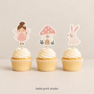Może przedstawiać: Trzy babeczki z ozdobnymi papierowymi topperami na drewnianych patyczkach. Toppery przedstawiają wróżkę o brązowych włosach w różowej sukience, różowy domek w kształcie grzyba w białe kropki oraz jasnobrązowego królika. Poniżej znajduje się napis "bella print studio".