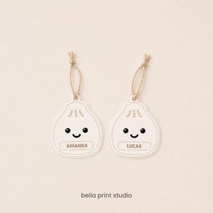 May include: Two white, flat, dumpling-shaped ornaments with a smiling face graphic, each featuring a tan string loop at the top. One ornament is personalised with the name AMANDA and the other with LUCAS. The text bella print studio is at the bottom.