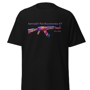 Puede incluir: Camiseta negra con un gráfico colorido de un rifle de asalto AK-47. El diseño del rifle incorpora varios colores y patrones. El texto encima del rifle dice "Автомат Калашникова-47" con "Since 1949" debajo.