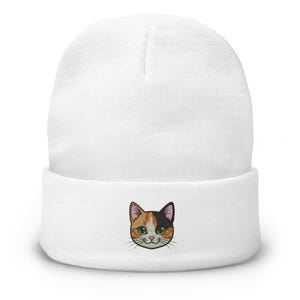 May include: A white beanie hat with an embroidered cat face. The cat face has orange, black, and white fur, green eyes, and a smiling expression. The beanie is made of a soft, knitted material.