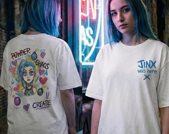 May include: White t-shirt with a cartoon illustration on the back of a blue-haired character, with the words "POWDER CHAOS CREATE". The front of the shirt has the text "JINX was here" in blue.