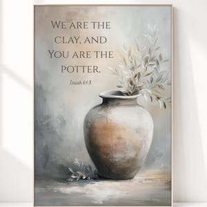 May include: A framed wall art print featuring a neutral-toned ceramic vase with a gold-colored crack detail, holding leafy branches. The text reads: WE ARE THE CLAY, AND YOU ARE THE POTTER. Isaiah 64:8. The background is a soft, blended gray and beige.