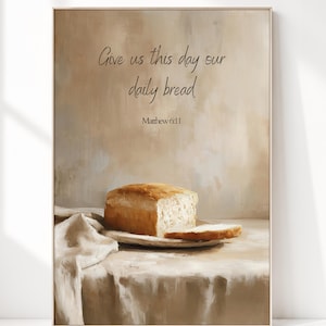 Op de afbeelding: Een ingelijste wandkunstprint met een brood op een bord met een stoffen servet. De tekst luidt: Give us this day our daily bread Matthew 6:11. De achtergrond is een neutrale beige en bruine geschilderde textuur met zichtbare penseelstreken.