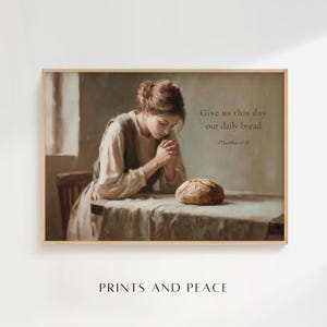 May include: A framed print featuring a woman with clasped hands, a loaf of bread, and the text "Give us this day our daily bread" from Matthew 6:11. The artwork has a vintage, painterly style with soft lighting and neutral tones.