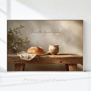 Può includere: Una stampa murale rettangolare che mostra una panca in legno rustico con una pagnotta rotonda su un panno bianco e una tazza in ceramica. Il testo "Give us this day our daily bread" è centrato sopra il pane. Un ramo d'ulivo è sulla sinistra.