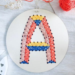 Könnte beinhalten: Ein handgestickter Buchstabe "A" auf einem weißen, runden Canvas. Die Stickerei verwendet rosa, rote, gelbe und blaue Fäden, akzentuiert mit schwarzen Perlen. Nadel und orangefarbenes Garn deuten auf ein Bastelprojekt hin.